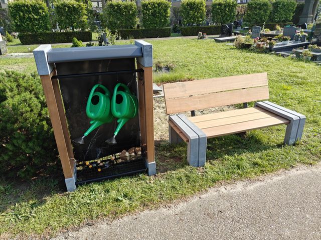 Maatwerk gieterrek van hout en beton met groene gieters en houten bank op een begraafplaats